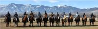 A row of riders faces the camera. All wear light blue denim shirts and helmets. There is a desert mountain range under a blue sky in the background.