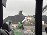 View from a vehicle showing heavy machinery and large coal piles at an industrial site, with conveyor belts and cloudy sky.