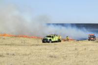 Firefighters in yellow gear monitor a controlled grass fire spreading across a dry field under a clear sky.