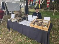 Outdoor display table with antique diving helmet, boots, weights, nautical tools, maps, and informational signs at a historical event.
