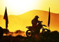 A dirt bike rider is silhouetted against a low sun over some hills in the distance. Two flags mark the race course.