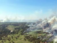 Aerial view of a large forest fire spreading smoke and flames across a green landscape under a clear blue sky.
