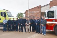 Firefighters in uniform stand in front of two large fire trucks one is green the other is red