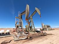Two large, olive-green industrial pumpjacks stand in a flat, arid desert landscape under a clear, bright blue sky. In the foreground and mid-ground, various storage tanks, white pressurized vessels, and insulated piping systems are visible on the sandy ground.