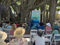 Artist painting a marine scene on a large canvas outdoors, with an audience seated on white chairs under large tree branches.