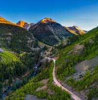 This photograph provides an overhead view of a curvy dirt road with an uneven surface as it winds through a mountainous landscape with an extreme cross-slope. Coniferous trees are growing thickly on either side of the road, and in the far distance several mountain peaks with patches of snow are lit by sunlight below a clear blue sky.  