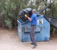 A man throws a bag of trash into a blue dumpster.