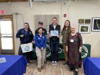 Group of six people standing indoors in front of tables with Federal Junior Duck Stamp banners, one person holding a duck painting.