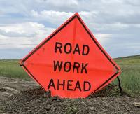 Bright orange diamond-shaped sign reading 'ROAD WORK AHEAD' placed on a dirt road with grassy fields and cloudy sky in the background.