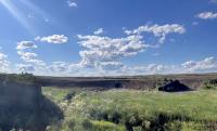 Culvert in a road traversing a wide open grassland under a bright blue sky filled with scattered white clouds on a sunny day.