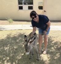 A woman wearing sunglasses smiles while petting a young donkey in a shaded outdoor area near a building.