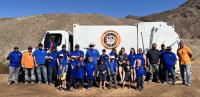 A Yuma County Public Works truck and a large roll-off dumpster sit in a desert cleanup area as volunteers collect trash across rocky public lands near Yuma, Arizona.