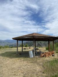 Covered wooden picnic shelter in an open grassy area with stacked firewood, a metal trash can, and distant forested mountains under a partly cloudy sky.