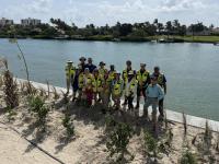 Group of people wearing safety vests standing on sandy shore near a body of water with greenery and buildings in the background.
