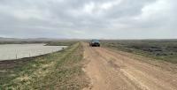 A pickup truck travels along a dirt road crossing an earthen dam amid a prairie landscape.