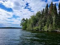 A scenic view of a forested shoreline under a partly cloudy sky, with calm lake water in the foreground.