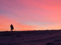 Photo of a pink sky in the background with a hunter silhouetted on the left hand side. Rifle stock is visible along with a dark landscape foreground. 