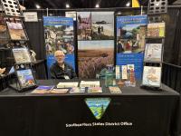 A person sits behind an informational booth for the Bureau of Land Management’s Eastern States office at a convention or expo. The table is covered with brochures, booklets, and educational materials. Large display banners behind the booth show outdoor scenes, wildlife, and public lands messaging. Two tall racks on either side hold additional publications. The person is seated at the center of the booth, smiling toward the camera.