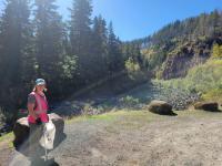 Person wearing a bright pink safety vest standing on a dirt path near large boulders, with a forested river canyon and steep rocky slopes in the background under a clear blue sky.