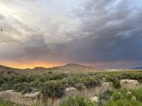 A dramatic desert landscape at sunset shows rugged hills and dense sagebrush in the foreground. The sky is filled with thick, layered storm clouds, shifting from deep gray to soft blue, while a bright band of orange light glows along the horizon, illuminating the mountains beneath it.