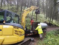 Workers using a yellow excavator to unload a large metal culvert from a trailer on a forest road.