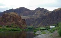 Riverbank campsite with two people sitting under a canopy beside a parked RV, set against steep rocky hills and a calm river.