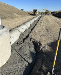 oncrete pipe sections set in fresh concrete along a dirt construction corridor, with an excavator and other heavy equipment working in the background under a clear blue sky.