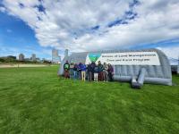 Group posing beside a Bureau of Land Management Cave and Karst Program tent, with a city skyline and cloudy blue sky behind.
