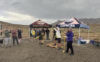 A couple dozen people stand around clapping while a person cuts a yellow ribbon; a cloudy desert landscape behind
