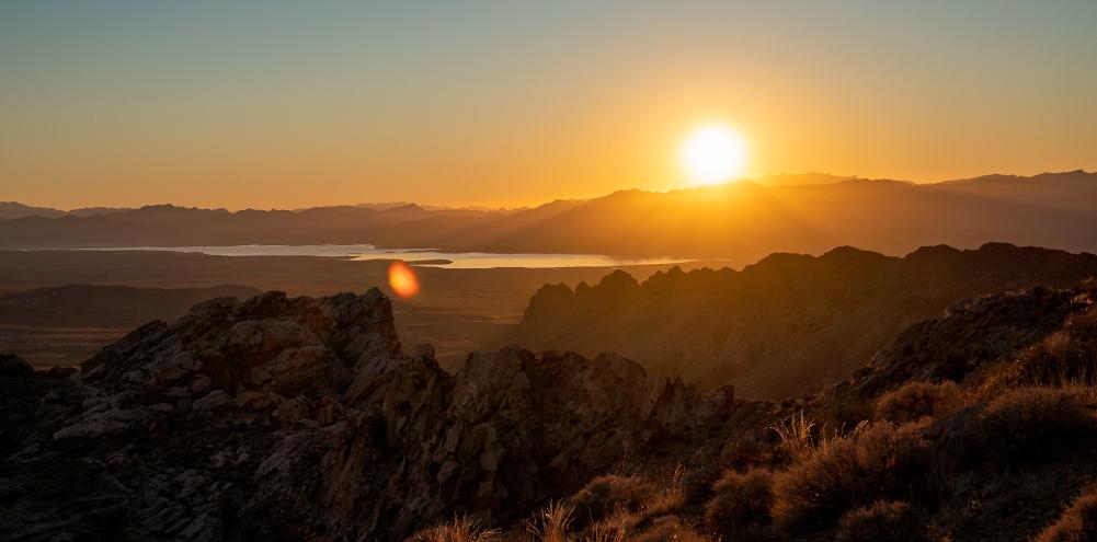 Sun rising just barely above the mountain range casting a bright orange and red light illuminating the valley, water and other mountains