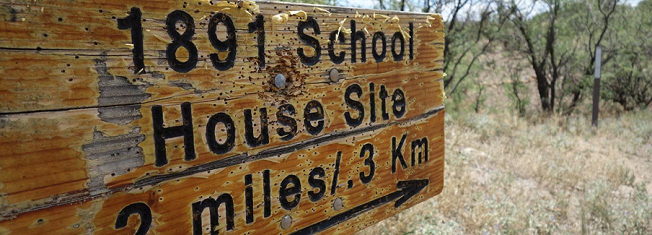 A wooden sign says, "1891 School House Site .2 miles/.3 km"