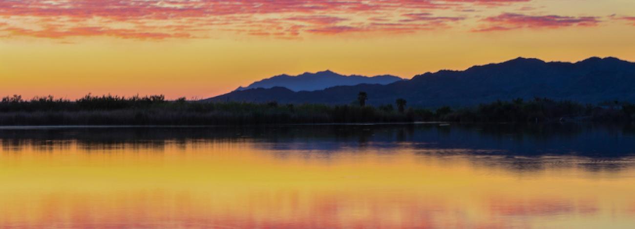 Calm lake reflecting a vibrant sunset sky with orange, pink, and blue hues behind distant mountain silhouettes.
