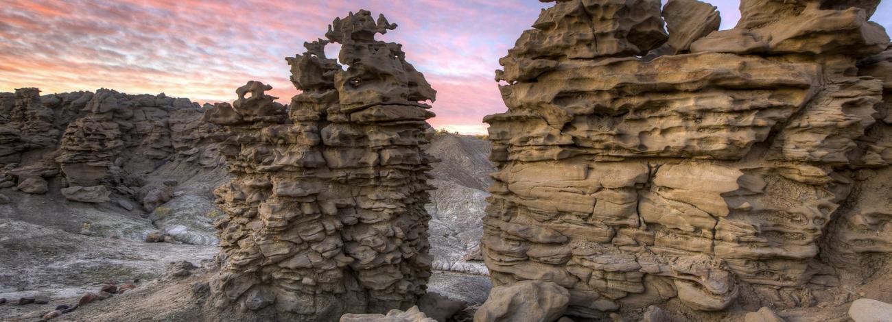 Brilliant hues of color fill the sky at sunset over rising pillars of sandstone that are worn away to reveal intricate patterns throughout with twisted knobs/ protrusions creating this fantasy like landscape.