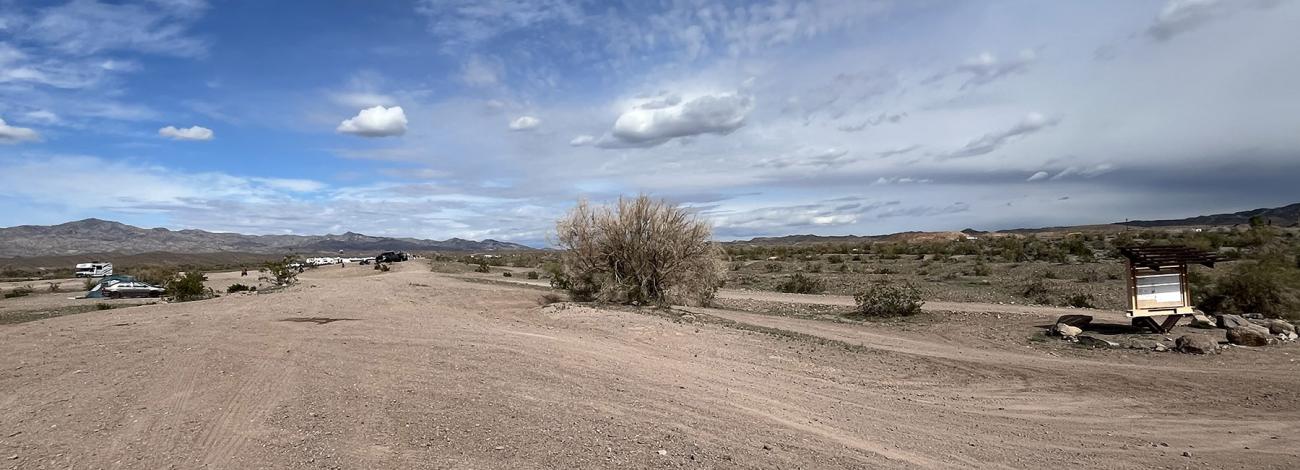 Wide dirt area stretching through a dry, open desert landscape under a partly cloudy blue sky.