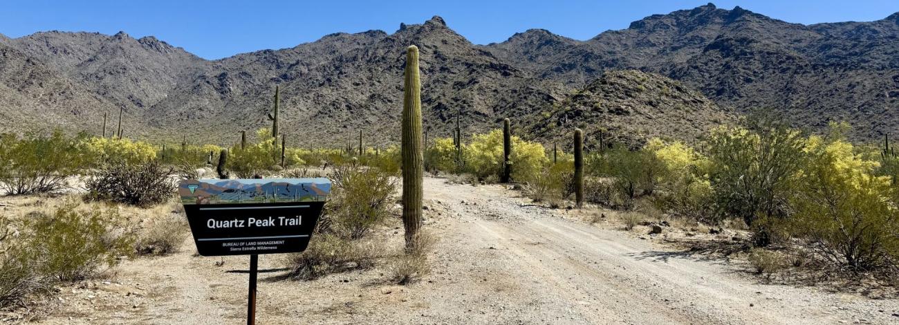 a road in a desert environment. A sign reads Quartz Peak Trail. A mountain range is in the distance.