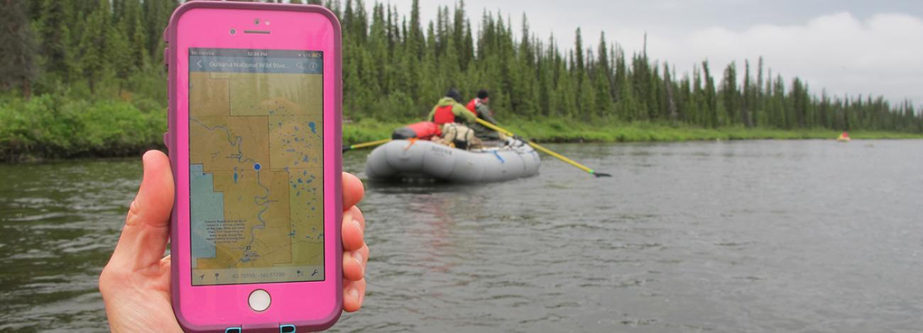 Rafter holding pink iPhone with Georeferenced PDF map displaying showing a blue dot of where they are located on a map. Fellow rafters shown ahead of them on the Gulkana River. 