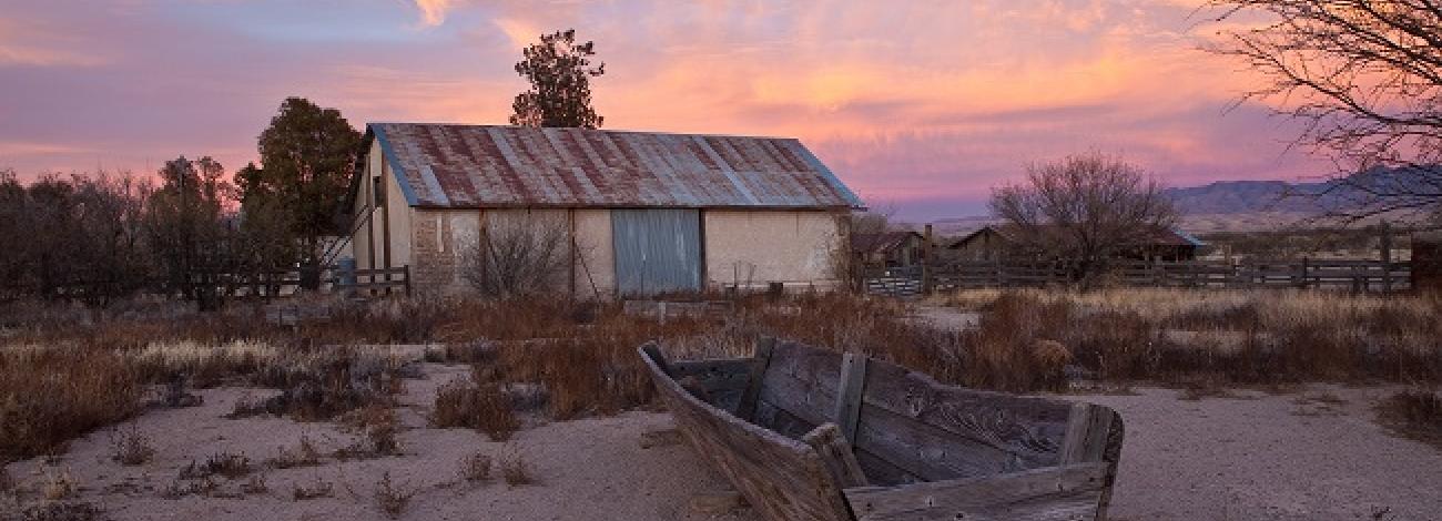 the wooden shell of an old boat in front of what appears to be an old barn which has a rusty corrugated tin roof.