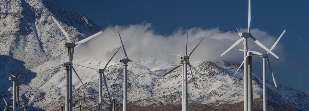 Image shows wind turbines creating energy by actively spinning in front of a background of rocky mountain under a cloudy sky.