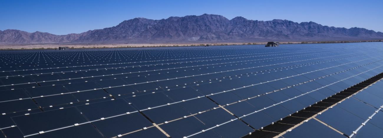 Image shows numerous rows of solar panels in the middle of a desert field surrounded by mountains under a clear sky.