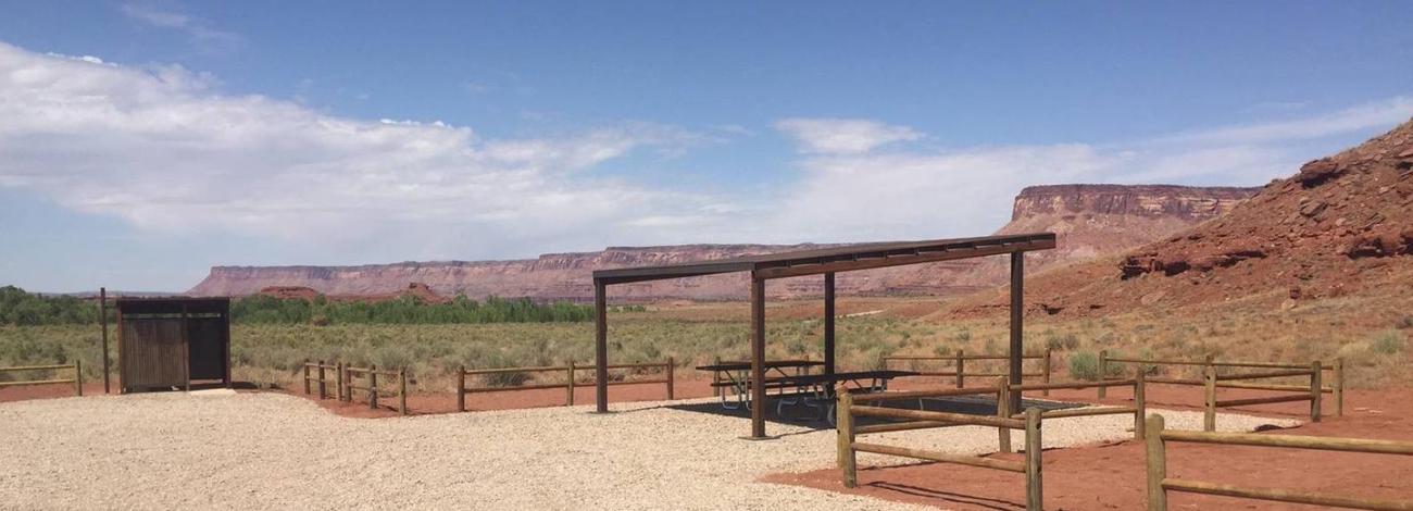 Superbowl group site shad structure and vault toilet with scenic red sandstone cliffs in background.