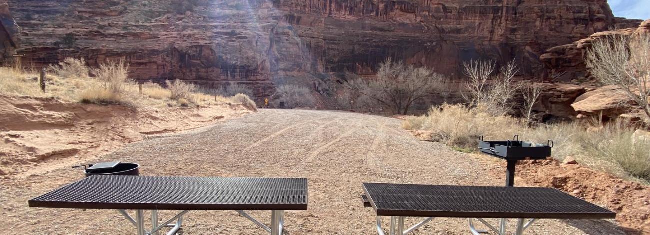 Group campsite, picnic tables, fire pit, and grill with sun setting behind cliffs in background
