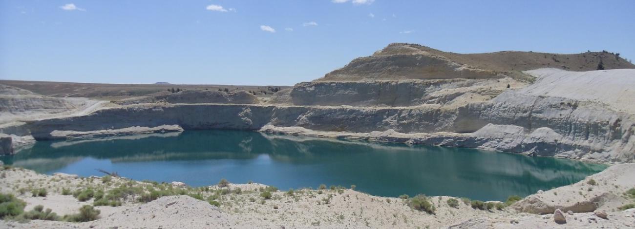 Image shows a body of water surrounded by mountain like hills in a desert under a cloudy blue sky. 