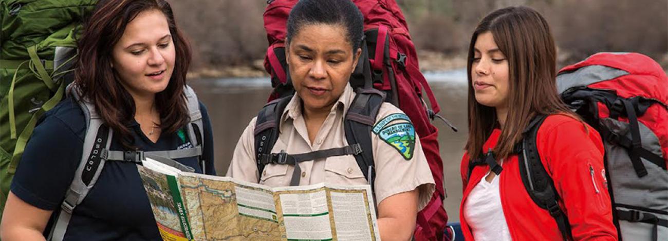 two hikers with backpacks stand on either side of BLM employee who is looking and pointing at a map they are holding.