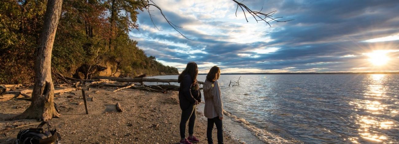 Two hikers looking out across the Potomac Heritage Trail and river at Douglas Point in Maryland with the run shining on the water. Photo by Bob Wick.