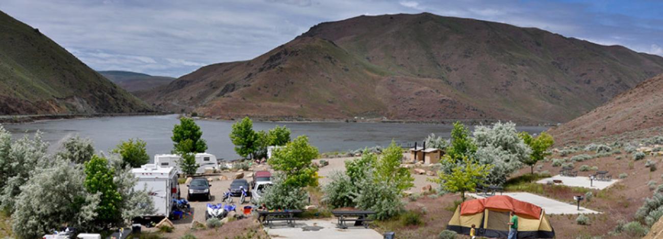 A campgroup along a lake in Idaho with concrete bases for tents, RVs, camp chairs, and tents scattered through the sites with trees spread throughout. A pit toilet and other amenities are also onsite.