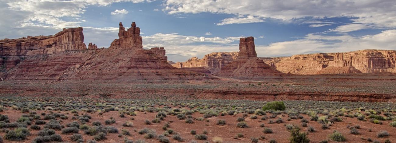 Red sandstone spires reach toward a cloud-filled blue sky across a barren desert.