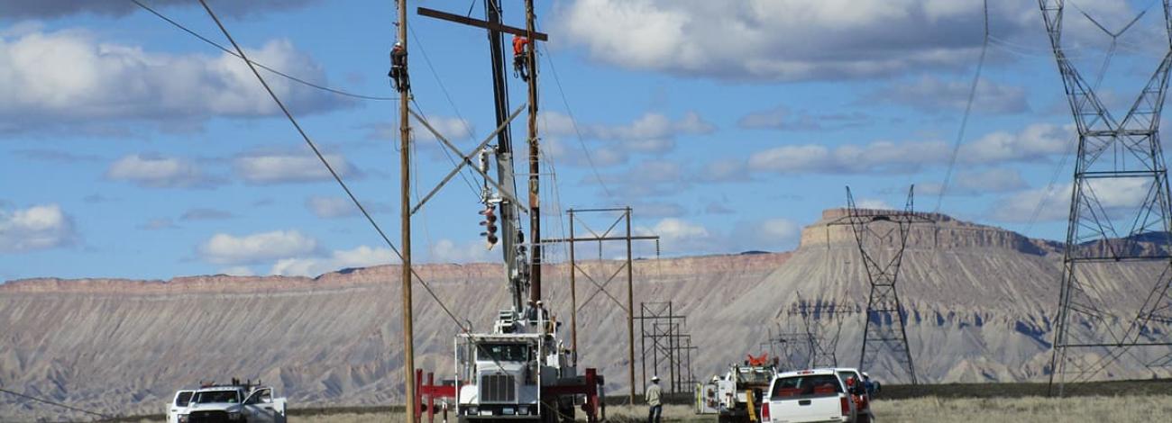 Cloudy blue sky with mountains in the background of men installing a powerline with two white pick-up trucks along with a utility truck installing a distribution power line with another Christmas tree power line to the right.