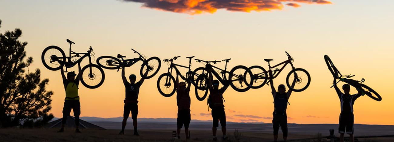 Six e-bike riders are silhouetted against a sunset as they all hold their bikes above their heads