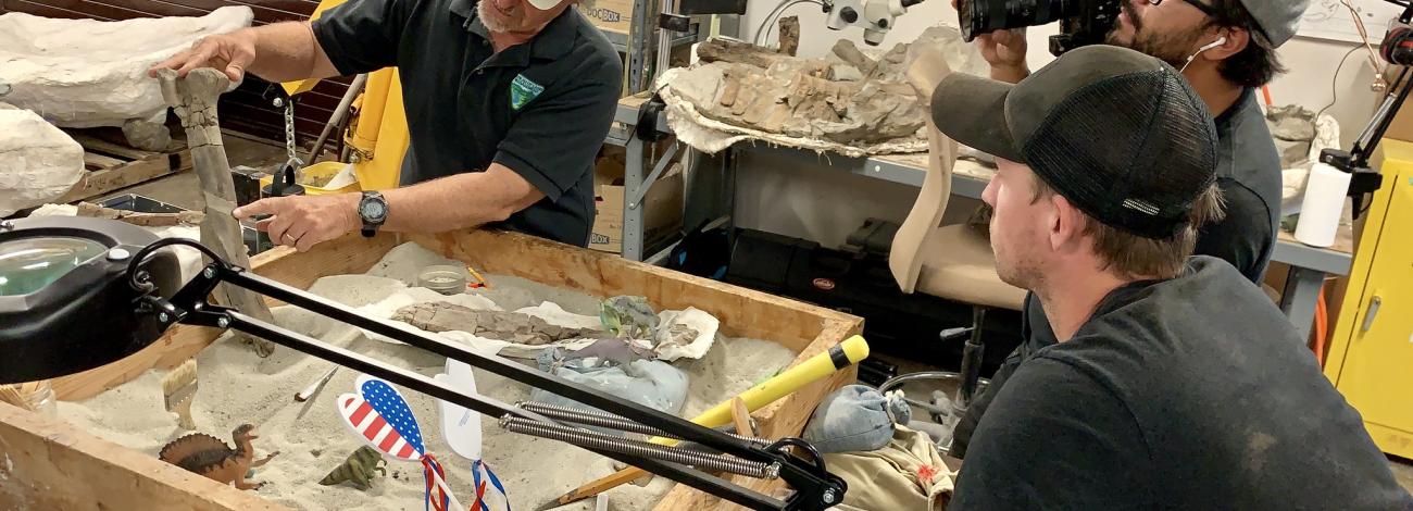 Paleontologist Dr. Titus points to a fossilized bone. Two journalists film him as he presents. 