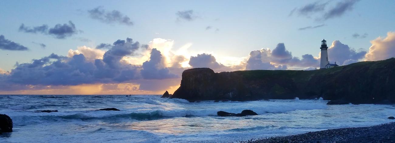 The Yaquina Head headland jutting out into the sea with the Yaquina Lighthouse on top. The sun is setting and the light is a deep blue.